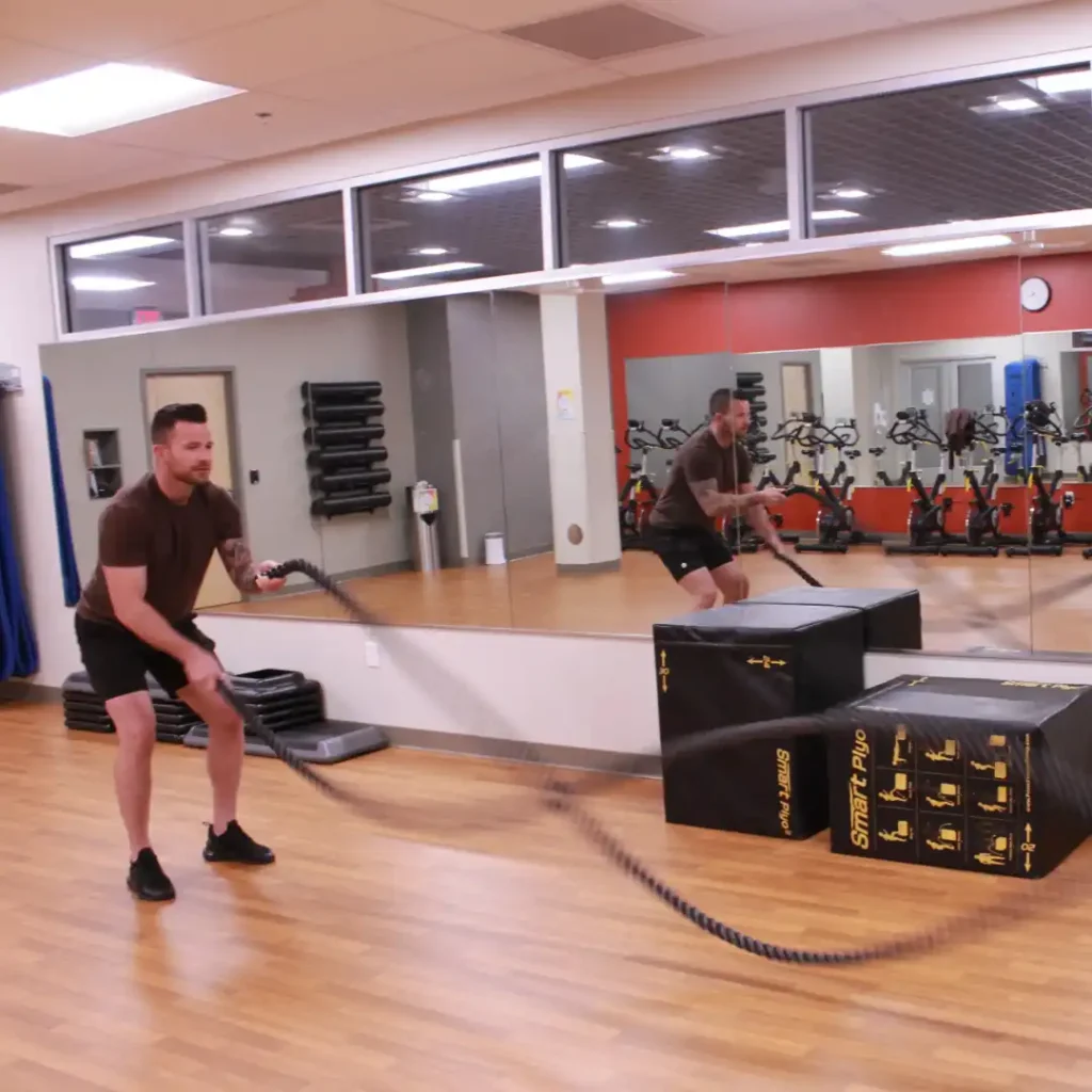 Good looking person engaged in a corporate fitness workout, vigorously swinging battle ropes. Their determined expression and powerful movements demonstrate intensity and focus, while the ropes create dynamic waves, emphasizing the high-energy of the exercise.