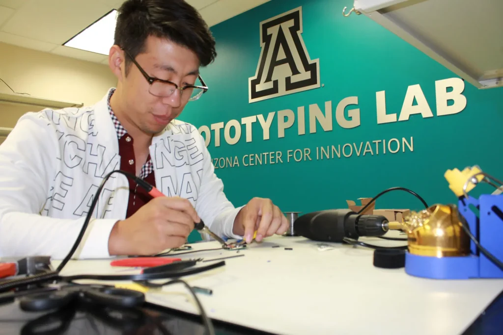 Entrepreneur at UA Tech Park, focused and intently soldering a prototype together, surrounded by tools and components, with a determined expression on their face. The room is washed in a calming teal backdrop, enhancing the creative atmosphere.