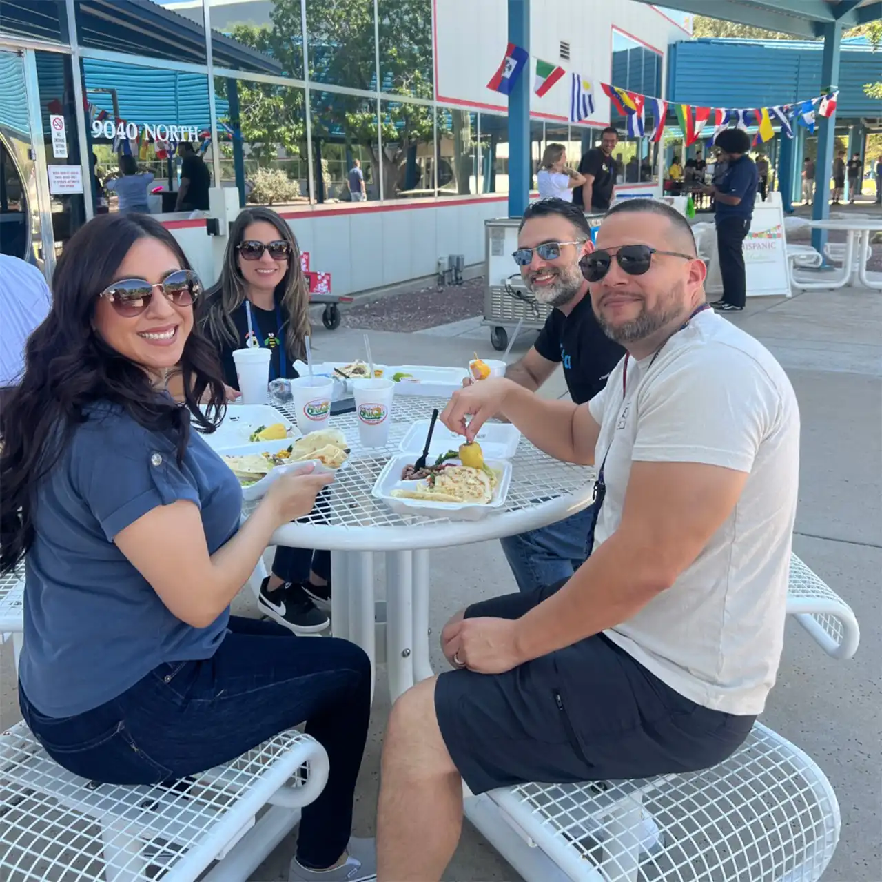 Employees gather outdoors, joyfully sampling various salsas and debating which is the tastiest, their smiles and laughter creating a lively atmosphere with Latin American flags waving proudly in the background