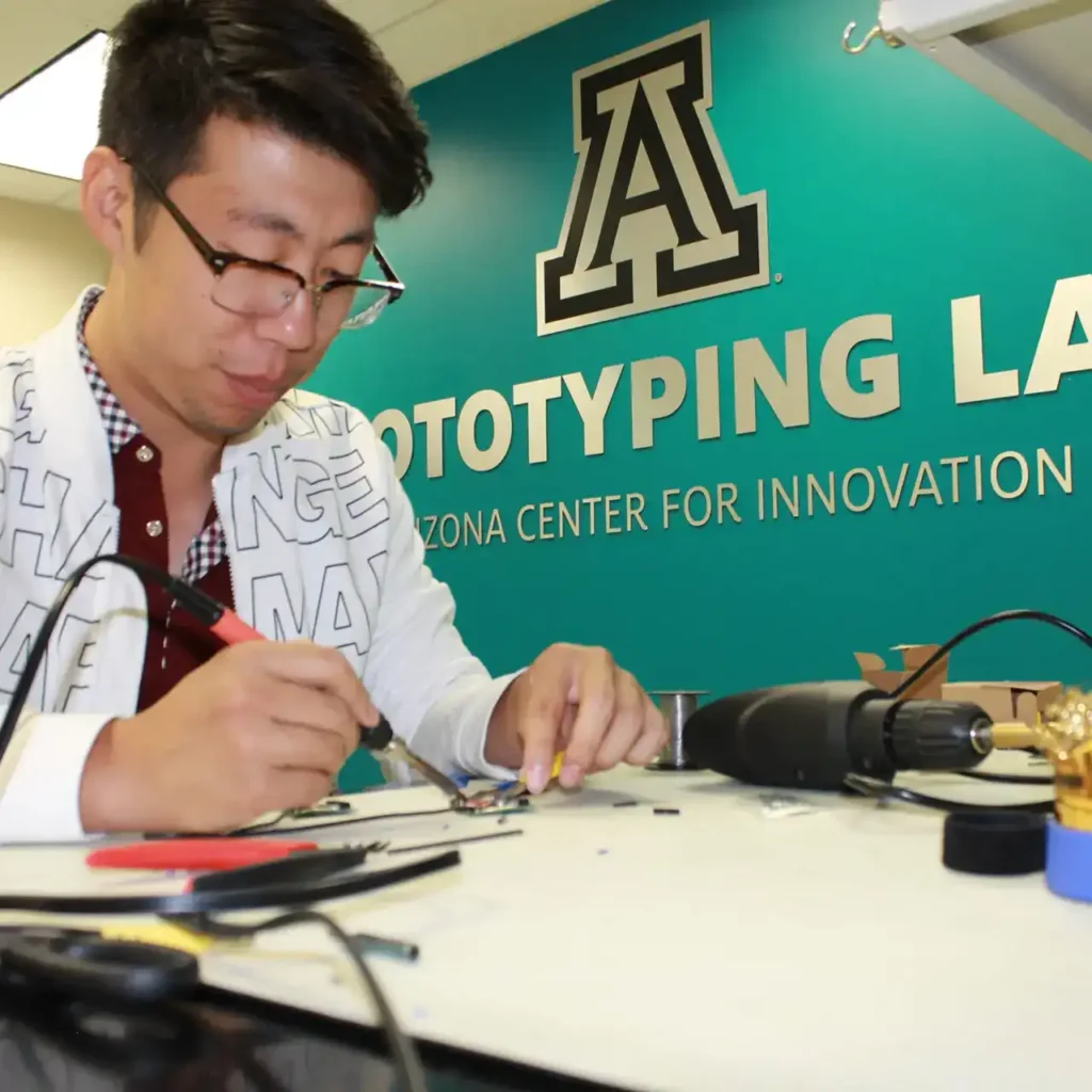 Entrepreneur at UA Tech Park, focused and intently soldering a prototype together, surrounded by tools and components, with a determined expression on their face. The room is washed in a calming teal backdrop, enhancing the creative atmosphere.