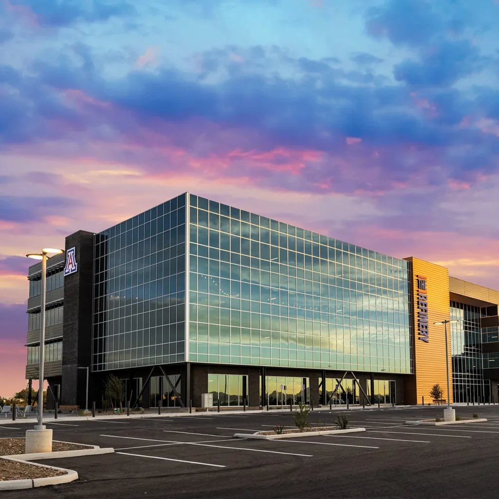 Modern glass building labeled “The Refinery” with a University of Arizona logo, set against a vibrant sunset sky and an empty parking lot.