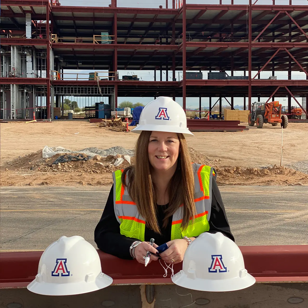 Woman smiling in a yellow construction vest wearing a white hard hat in front of a construction site with two hats on either side of her.