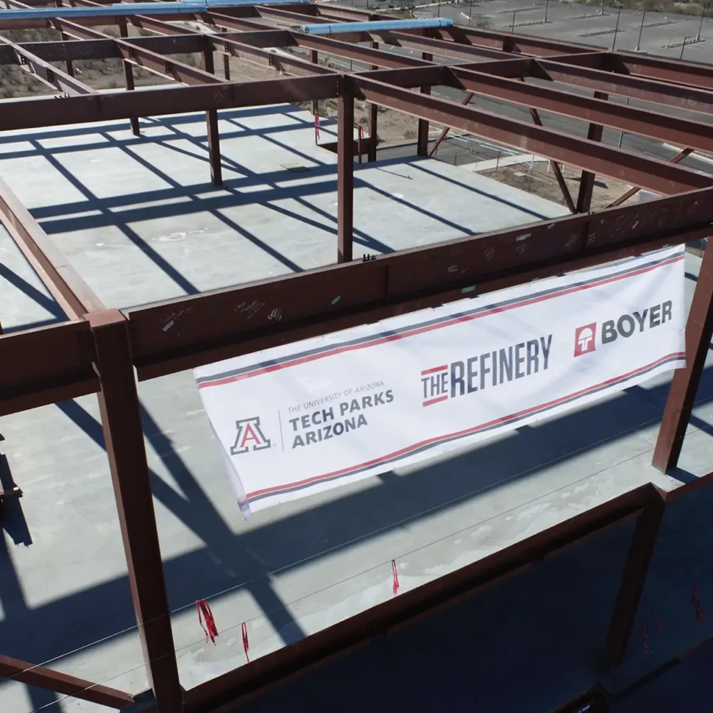 Construction beams with white banner hanging with three logos: Tech Parks Arizona, The Refinery, and Boyer.