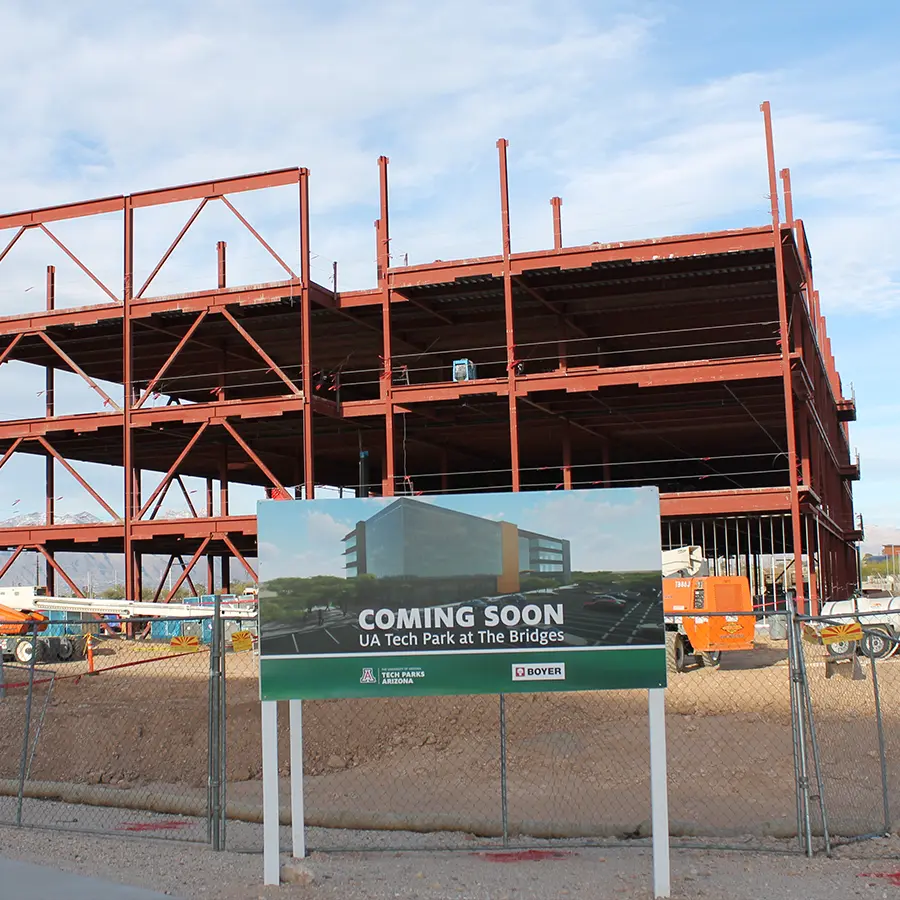 Steel-framed building under construction with a sign in front reading “Coming Soon: UA Tech Park at The Bridges.”