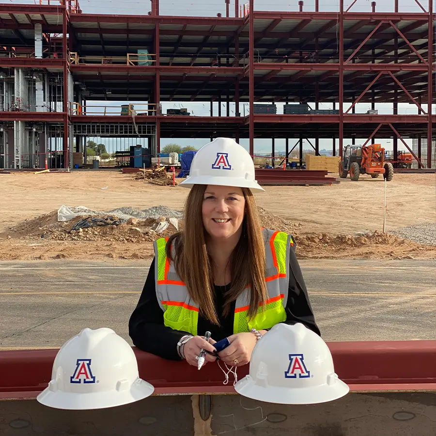 Woman smiling in a yellow construction vest wearing a white hard hat in front of a construction site with two hats on either side of her.