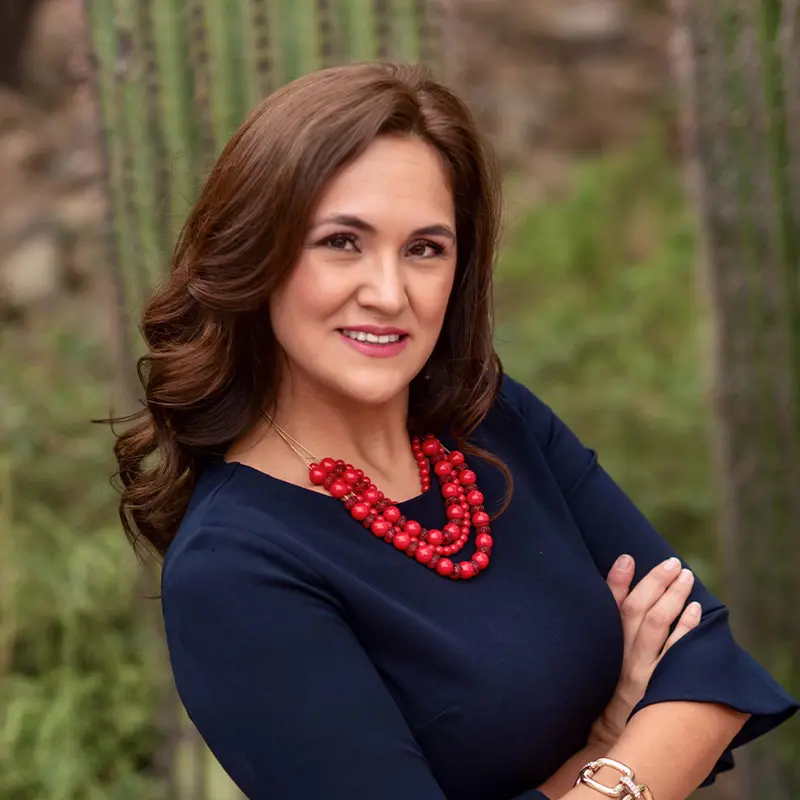 Headshot of Karla Morales wearing navy blue blouse and red beaded necklaces, crossing her arms in front of desert plants.