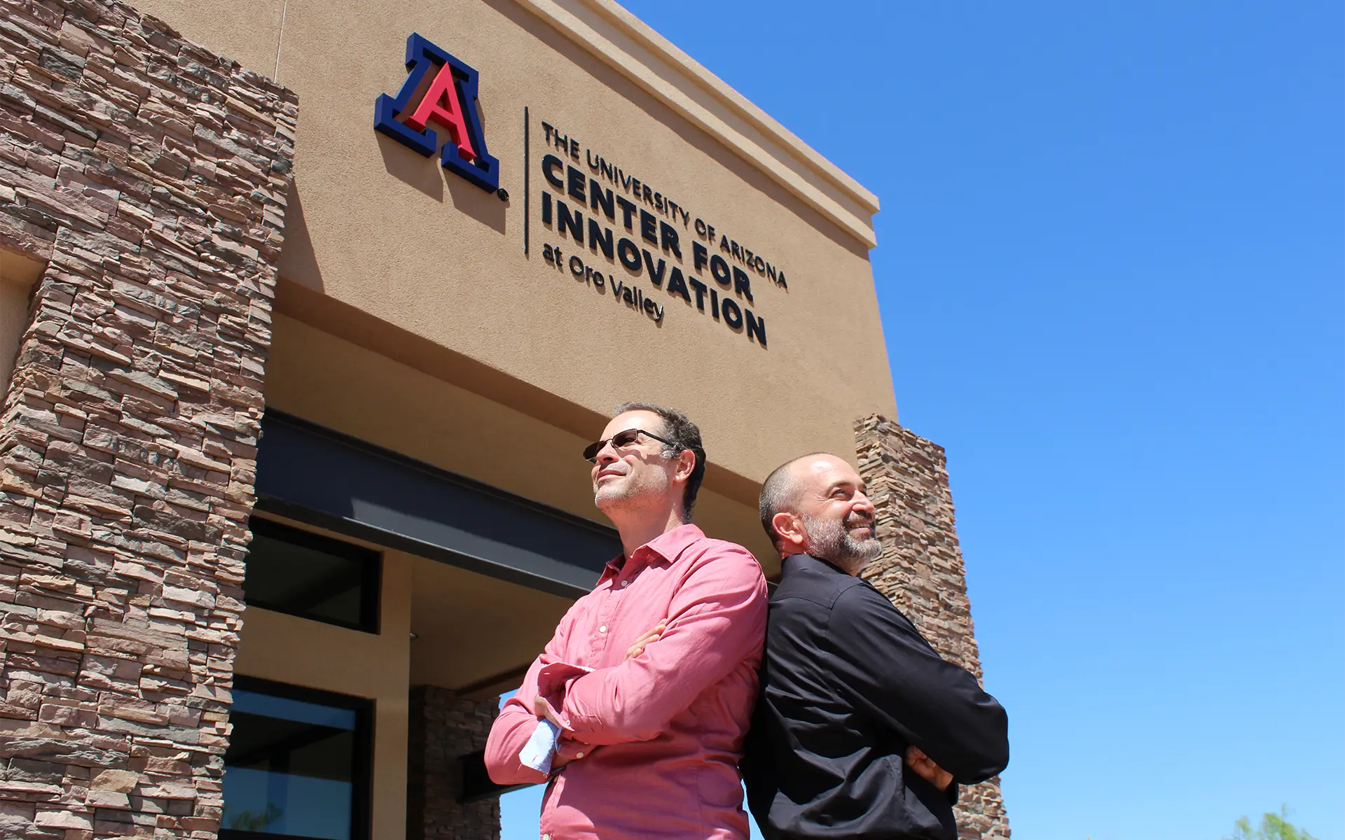 Two men standing back to back with their arms crossed in front of a Center for Innovation building in Oro Valley.