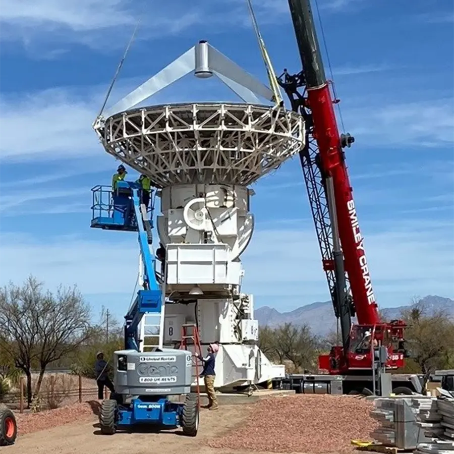 Workers use cranes and lifts to install or service a large white satellite dish antenna at a remote site with desert landscape and mountains in the background.