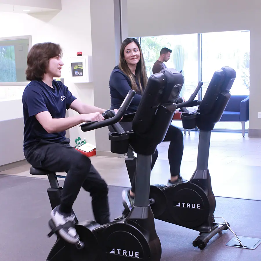 Two people talking while working out on stationary bikes.