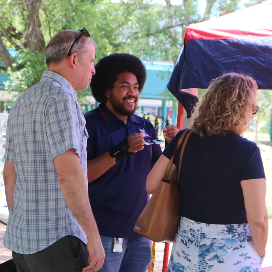 Three people chat and smile under a red and blue canopy at an outdoor event.