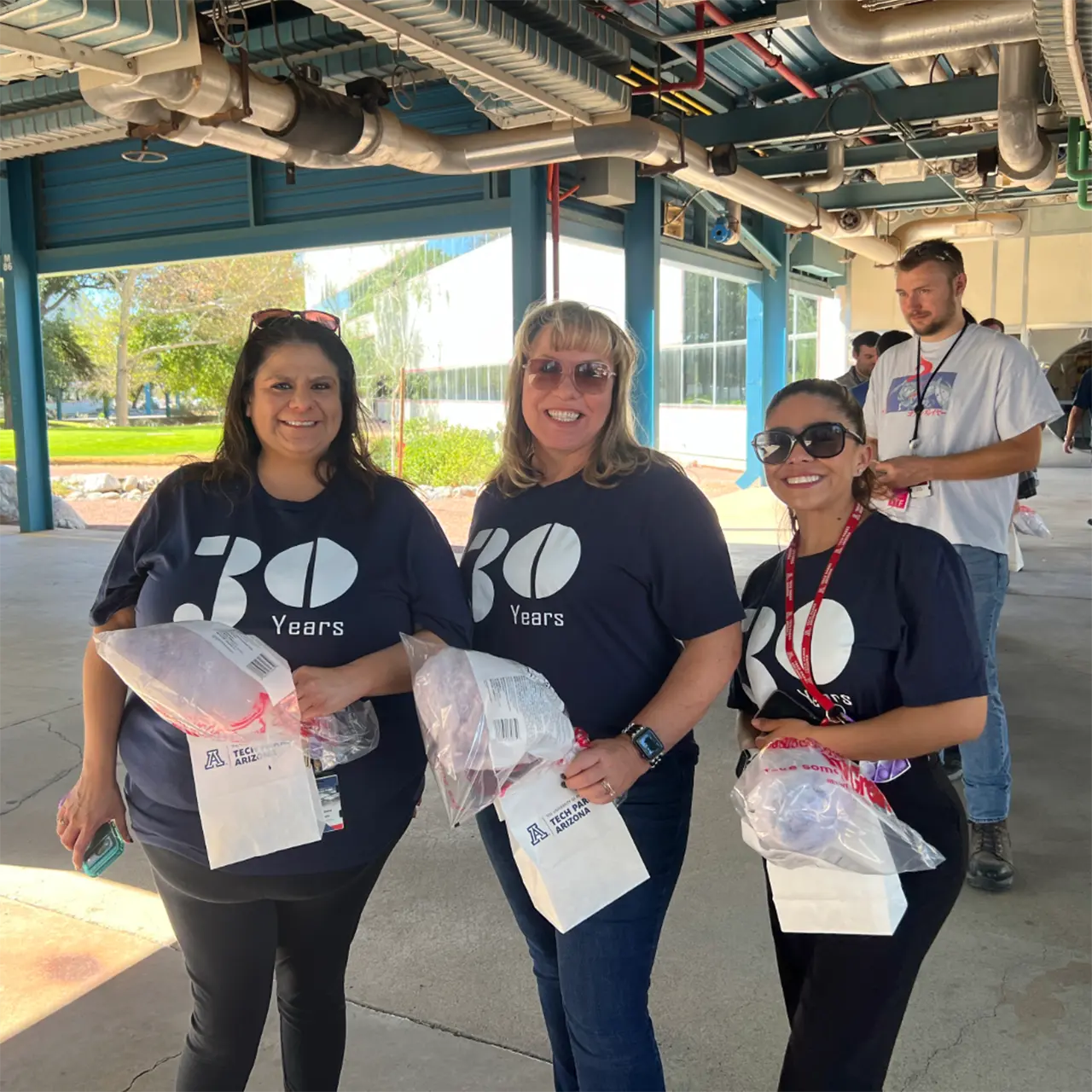 Three smiling women wearing "30 Years" shirts hold gift bags at an outdoor event.