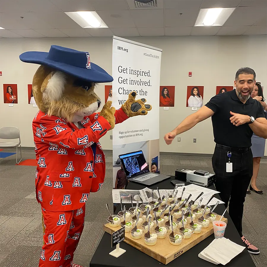 University of Arizona mascot Wilbur poses playfully with an event attendee near a dessert table at an indoor event.