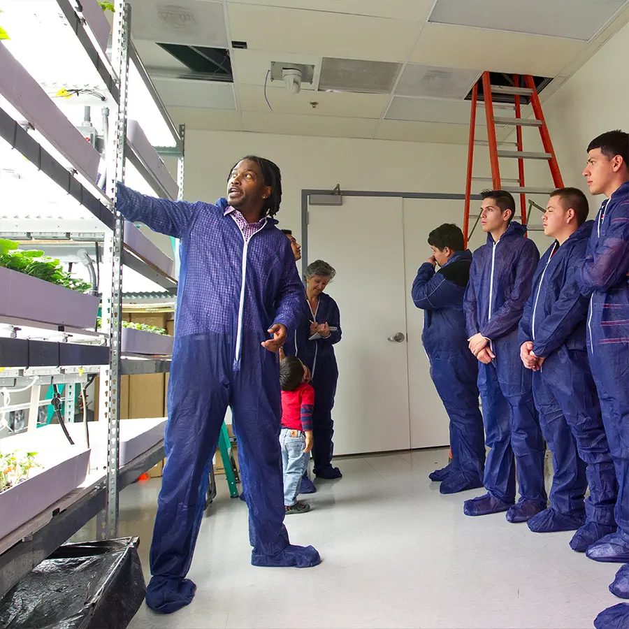 Instructor in a cleanroom suit explains vertical farming equipment to a group of students wearing matching suits during an indoor tour.