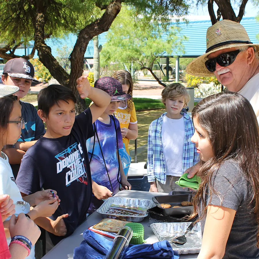Group of children gathered around an outdoor table with cooking trays, listening to an older man in a sun hat during a hands-on educational activity.