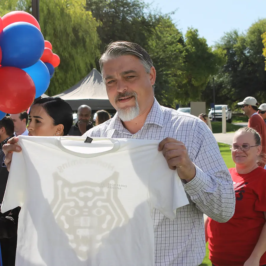 Older man posing for a photo holding up a white t-shirt with a Tech Cats logo on the front.
