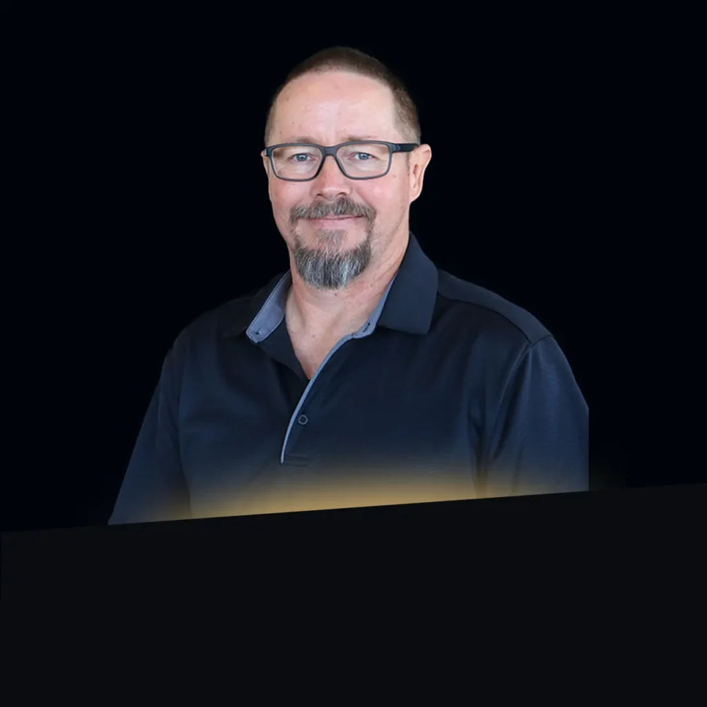 Man with glasses and goatee wearing a dark collared shirt, smiling against a black background.