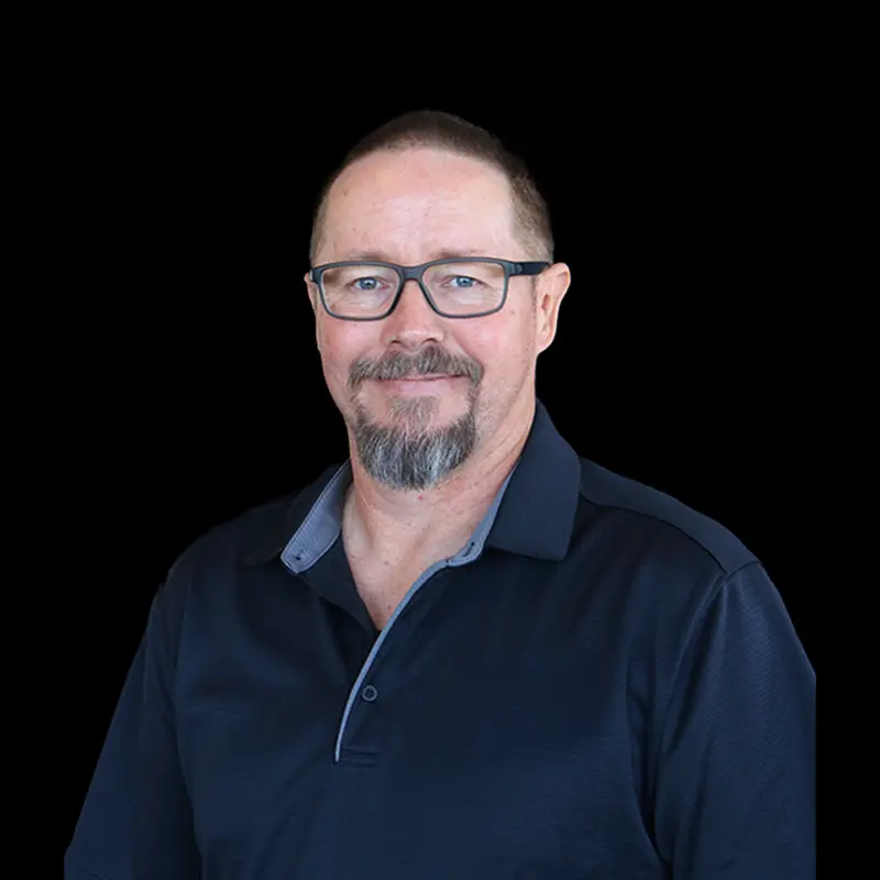 Man with glasses and goatee wearing a dark collared shirt, smiling against a black background.